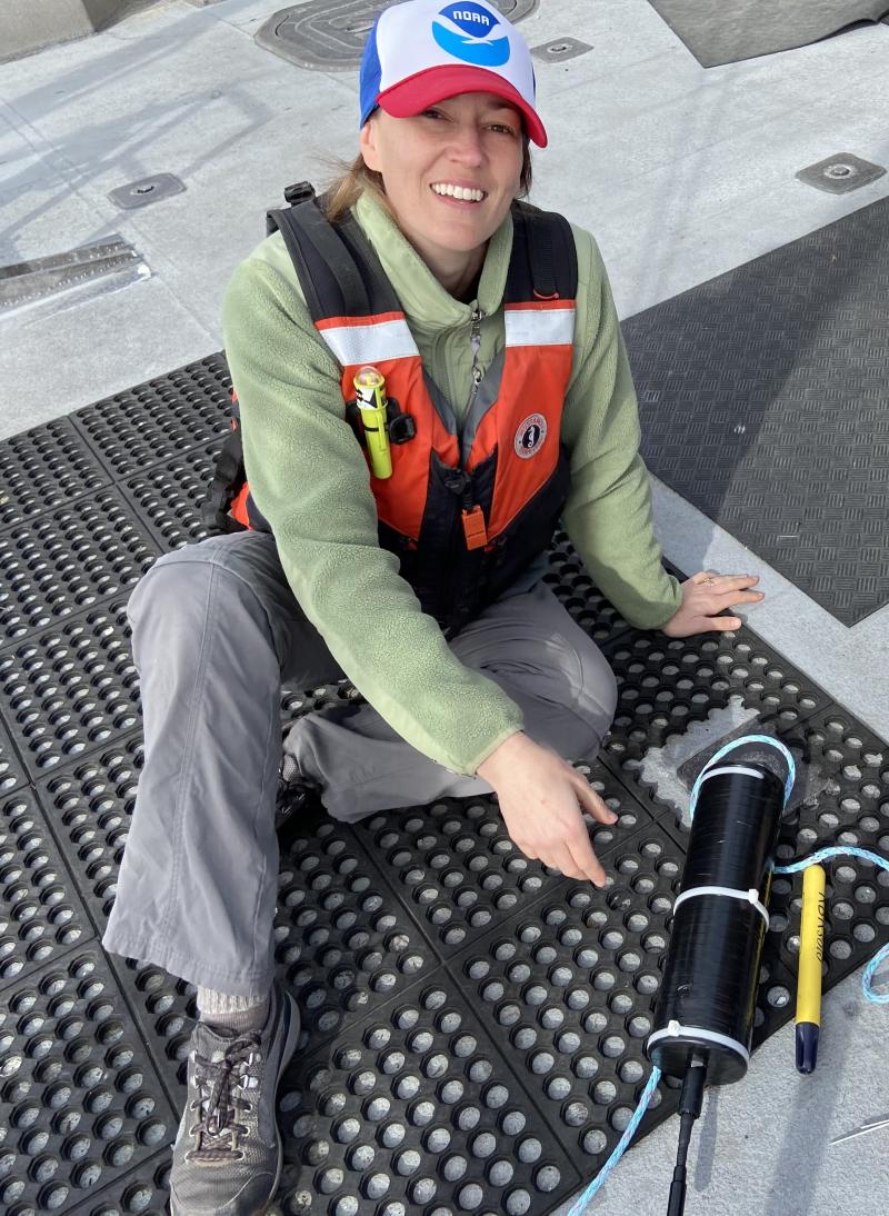 Lindsey Peavey Reeves sits on the back deck of the R/V Fulmar next to a SoundTrap underwater recorder. She looks up and is smiling at the camera.