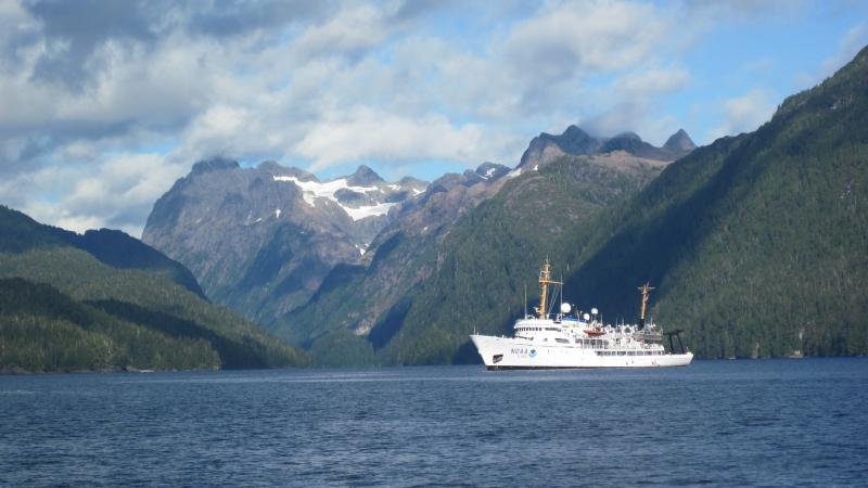 A large white ship with the NOAA insignia floats in a bay surrounded by mountains.