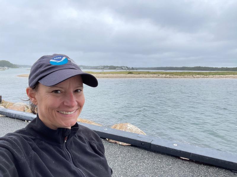 Woman scientist wearing NOAA hat and smiling stands in front of blue water with land and marsh grass in the distance.