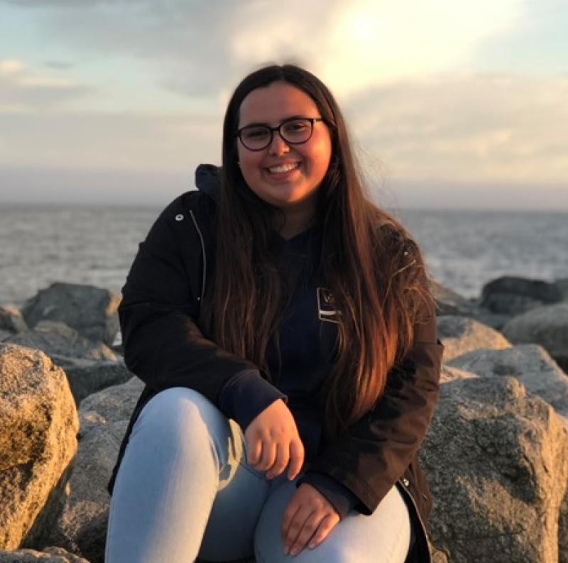Kourtney Burger sitting on the rocks with the ocean in the background. This is her bio photo and she is facing the camera and smiling