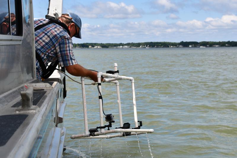 A man wearing a plaid shirt and a baseball cap leans out over the edge of a silver research vessel to retrieve a frame made of PVC pipe on which a camera is mounted.