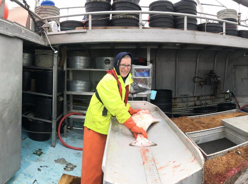 A woman in bright orange and yellow gear measures a large fish on the deck of a fishing vessel.