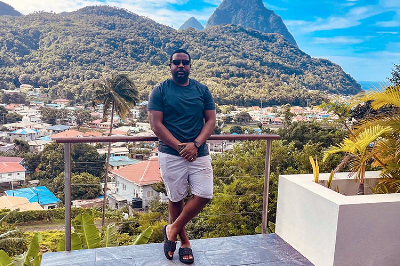 A black man wearing sunglasses, t-shirt, shorts, and sandals leans his back against a patio railing. In the background are tropical plants and palm trees, roof tops, and forested mountains.