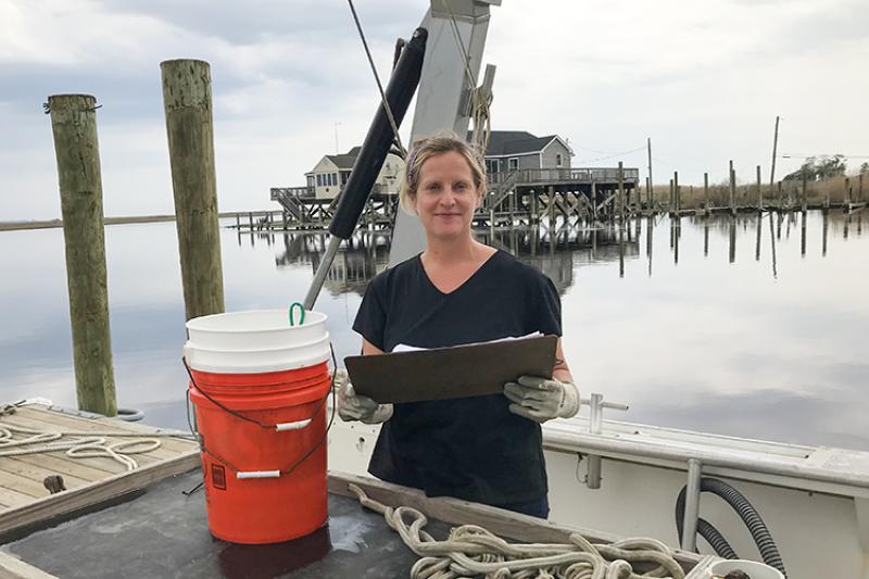 Ann Petersen wears heavy duty work gloves and holds a clipboard in her hand while standing on a docked boat. In front of her is a wood table that has some rope piled on it and to her right are two stacked plastic buckets. Behind her is a dock leading to a building built above the water on pilings.  