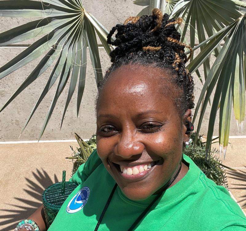 A woman in a green t-shirt smiles for the camera while standing in front of a concrete wall and leafy plants.