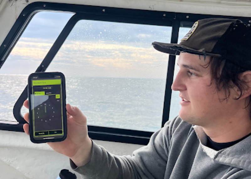 A man in a gray hoodie and hat holds up his phone to the camera while sitting inside a ship cabin.