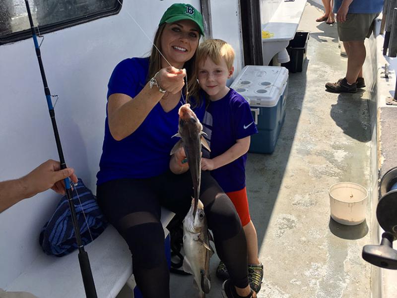 A white woman wearing a green hat and blue shirt sits on a boat next to a boy. She is holding a fish they caught.