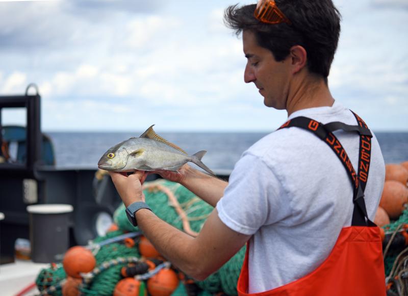 A man on the deck of a ship wearing orange overalls holds a silver fish and examines it.