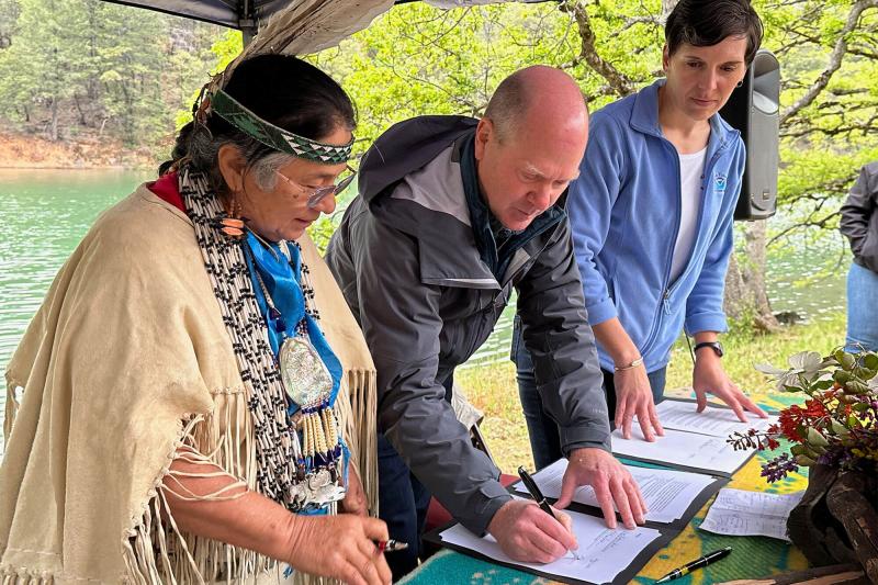 Three people signing documents on a table by a river bank