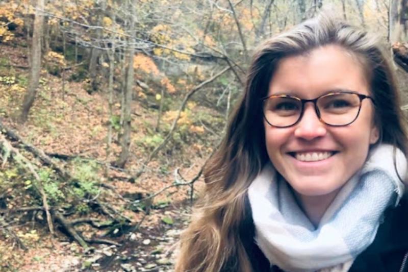 A woman with glasses, dressed warmly in a wooded area.
