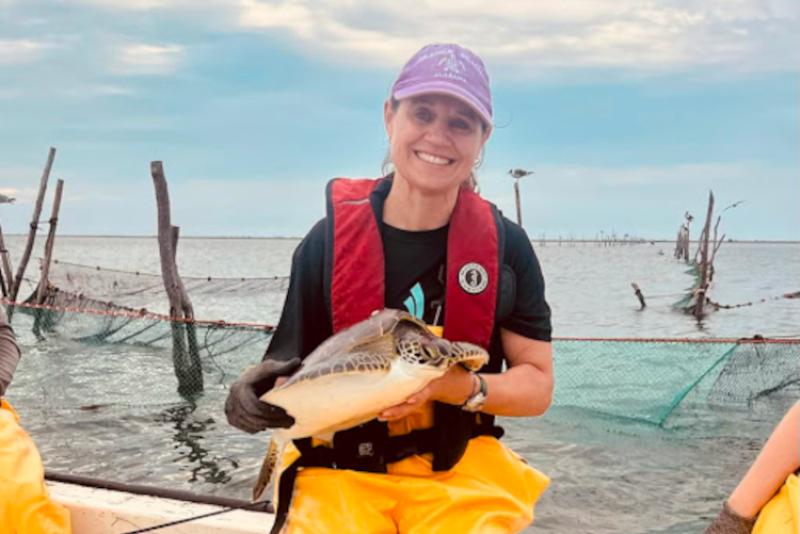A smiling woman sitting on the edge of a boat holding a small sea turtle.