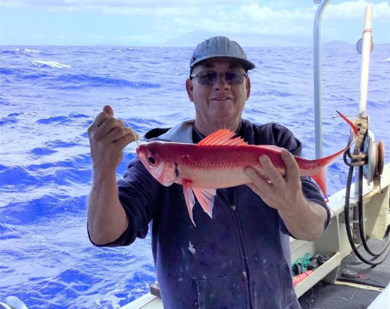 A fisher smiles while holding up a red and white fish, called an onaga, with both hands while out at sea.