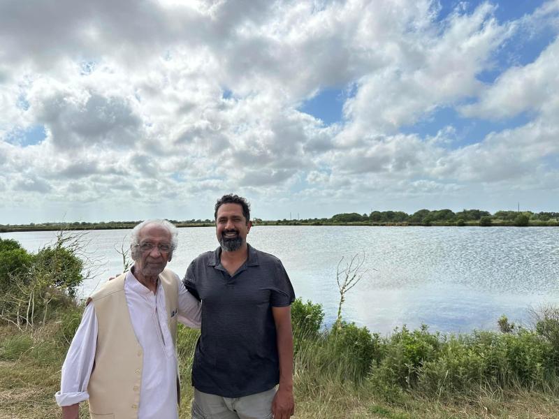 An adult man and his father stand with arms around each other with saltmarsh and a fish pond in the background. 
