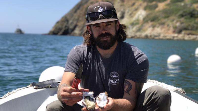 A bearded man wearing a hat on a boat holds a freshly shucked oyster.