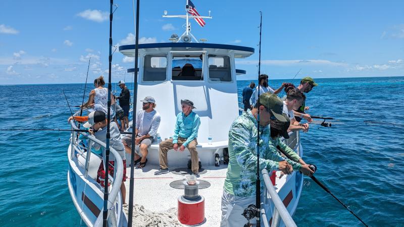 Roughly one dozen people, many wearing hats and active wear, stand along the railings of a white fishing boat, holding fishing poles.