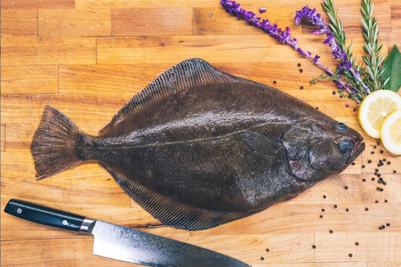 Petrale sole placed on a wooden counter next to a kitchen knife and fruit and floral garnish