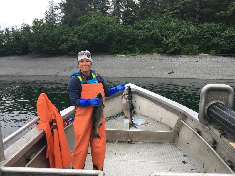 Teacher at Sea and Commercial Fisherwoman, Germaine Thomas, stands at the bow of her boat. She is dressed in orange coveralls, cold weather gear, and is holding two salmon--one in each hand.