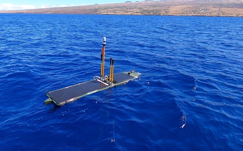 A rectangular autonomous surface vehicle floats on the ocean’s surface off the coast of a Hawai‘i shoreline.