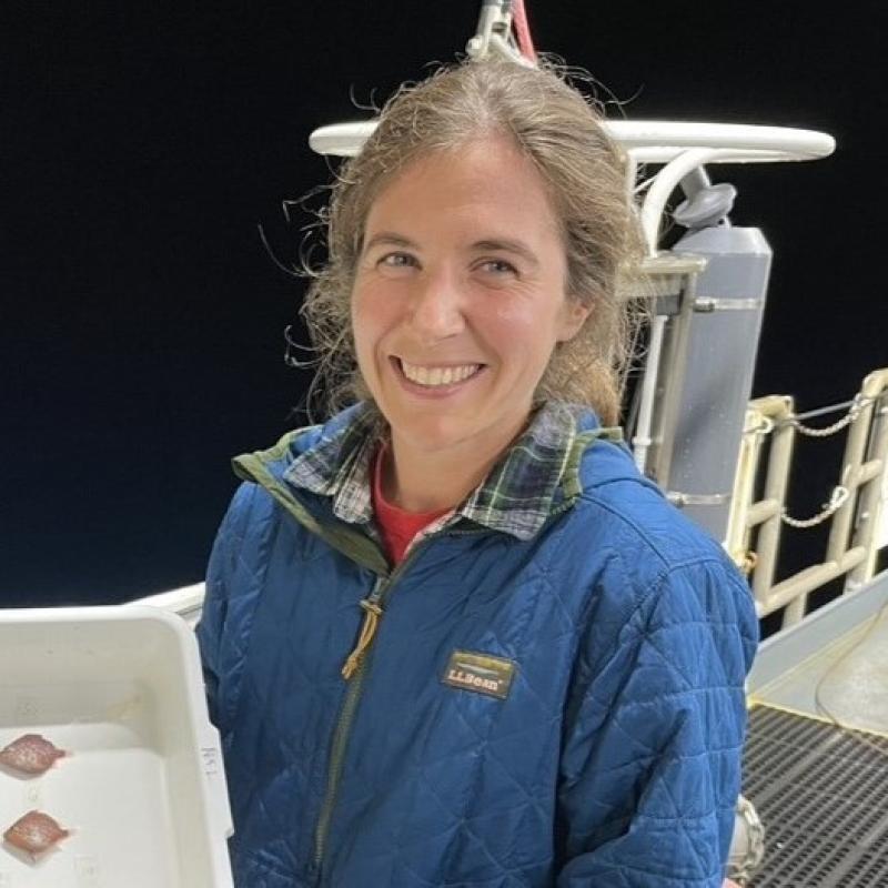 A woman in a blue jacket holds up a tray with fish while smiling at the camera on the deck of a boat.