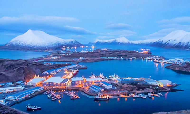 Aerial photograph of Dutch Harbor, Alaska. Snowcapped mountains surrounded by water and the lit up harbor town during the blue hour reflect creating a gradient between sea and sky. Credit: NOAA Fisheries