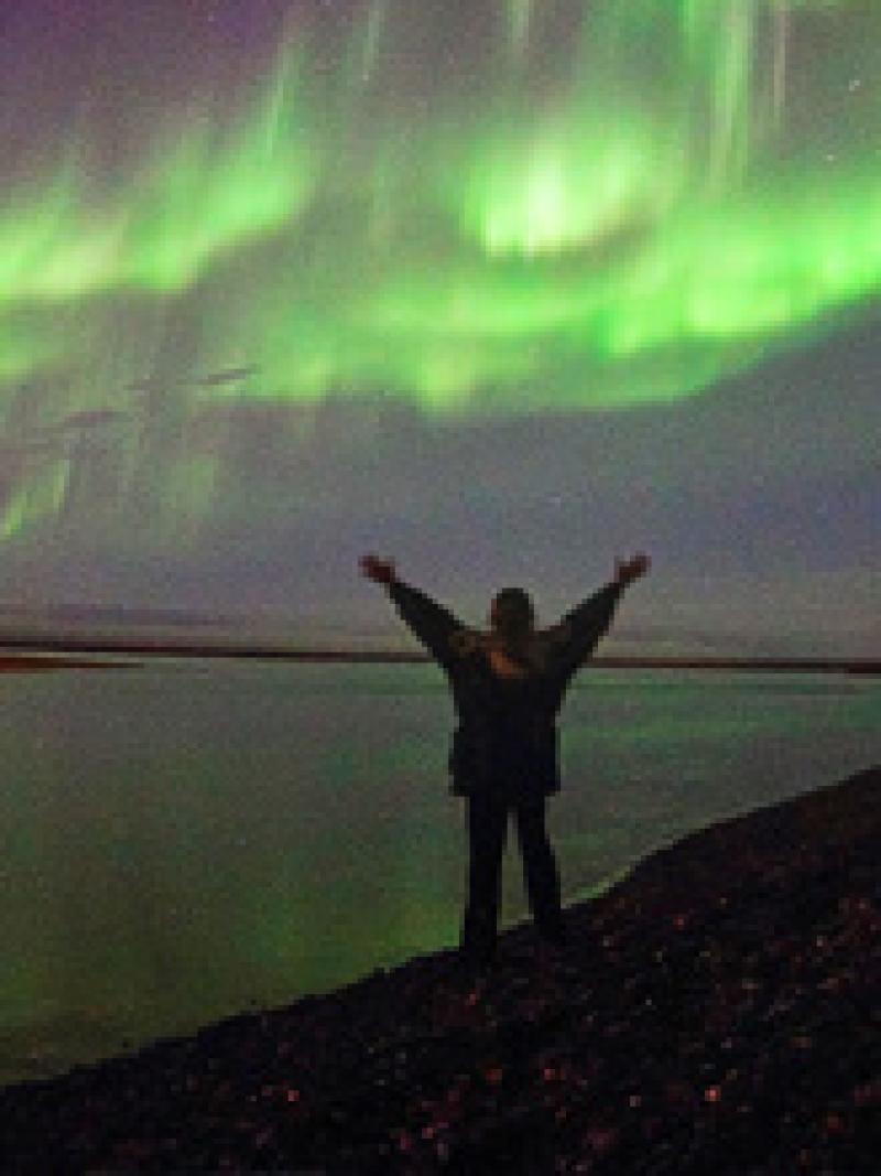 Woman with arms extended near the water with green lights in the sky 