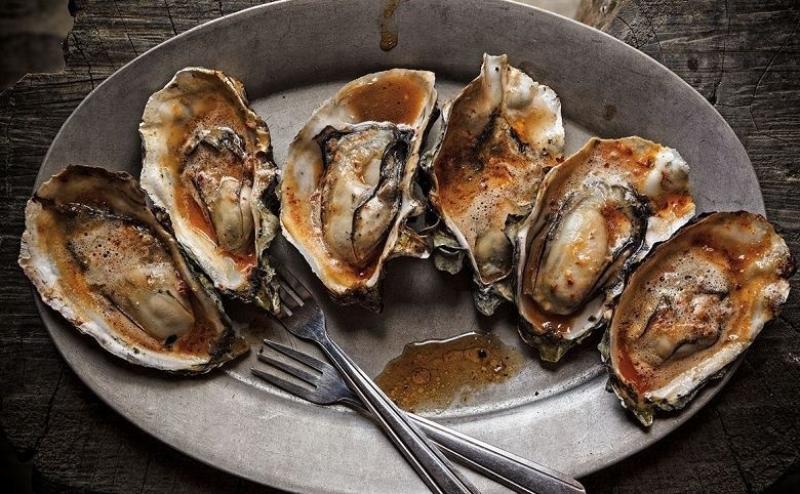 Six grilled Hog Island oysters drizzled in barbecue chipotle butter, on a serving dish with two small forks.