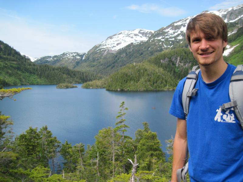 Brunette man wearing blue shirt and bookbag hiking near a lake with mountains in the distance