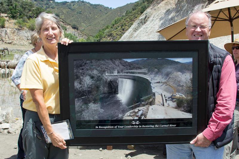 Two people holding a large, framed photograph of a dam