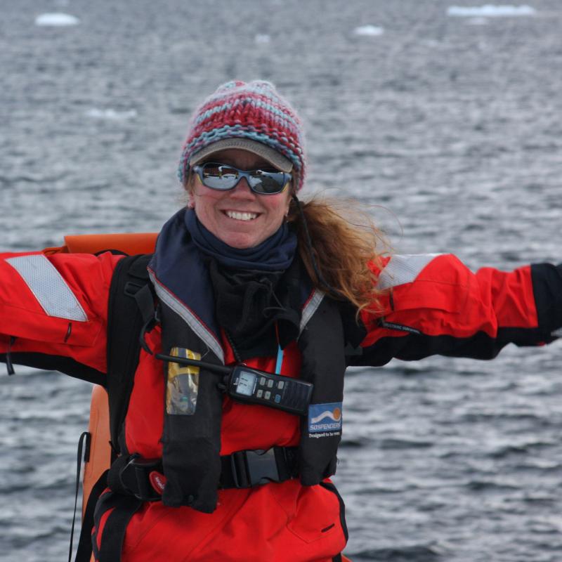 Woman wearing hat, glasses with orange coat with ocean and ice in background. 