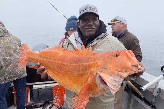 A bearded black man holds up a large, orange yelloweye rockfish using both hands and smiles for the camera. Three individuals stand behind him on a boat among fishing rods, buckets, caught fish, and other supplies.