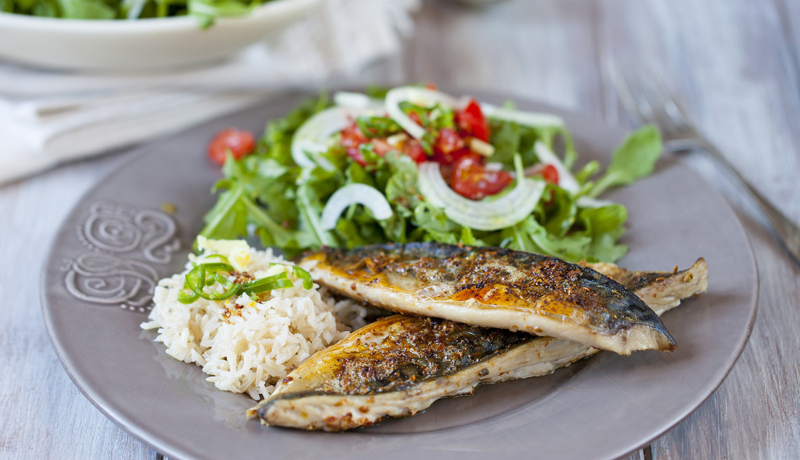 A blue-gray ceramic plate with two herb-broiled mackerel fish that have a buttery herb seasoning on them. There is also some salad and rice on the plate.